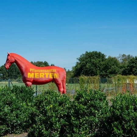 Eine große rote Pferdestatue mit der Aufschrift „Pflanzencenter MERTEN“ steht auf einer grünen Wiese.