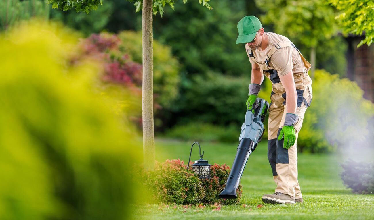 Eine Person in khakifarbener Uniform benutzt einen Laubbläser in einem üppigen Garten.